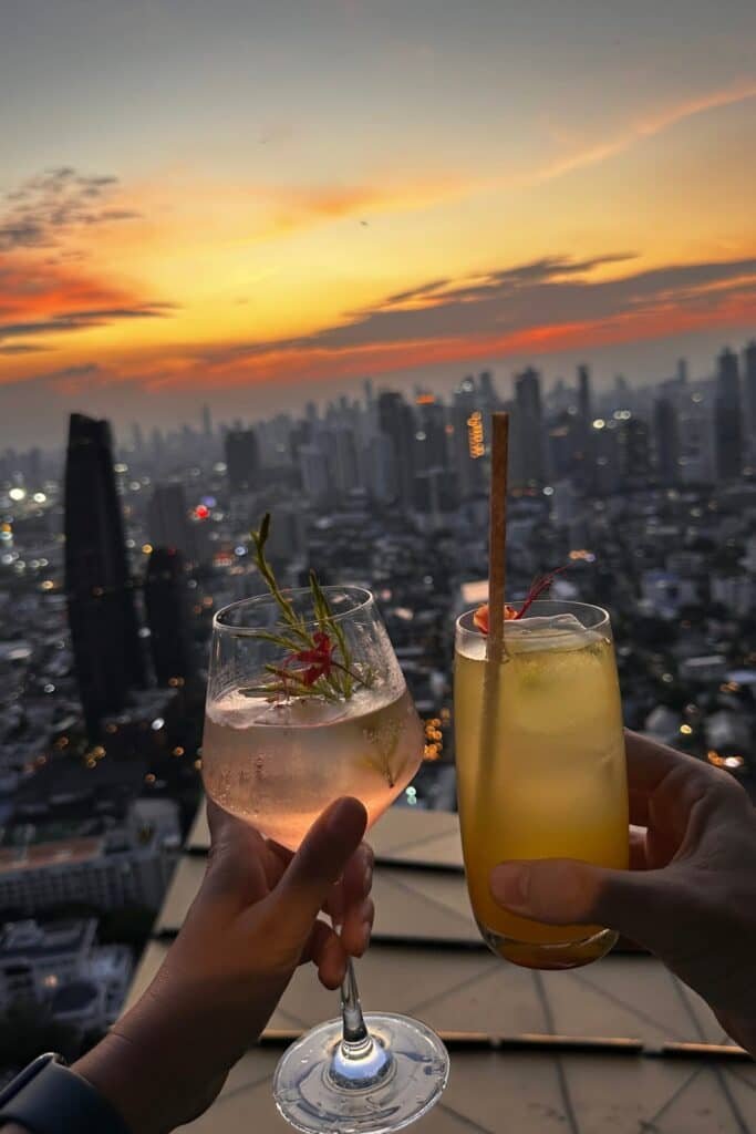 Bangkok skyline karşısında gün batımında rooftop barda kokteyl kadeh tokuşturma anı, Tayland.