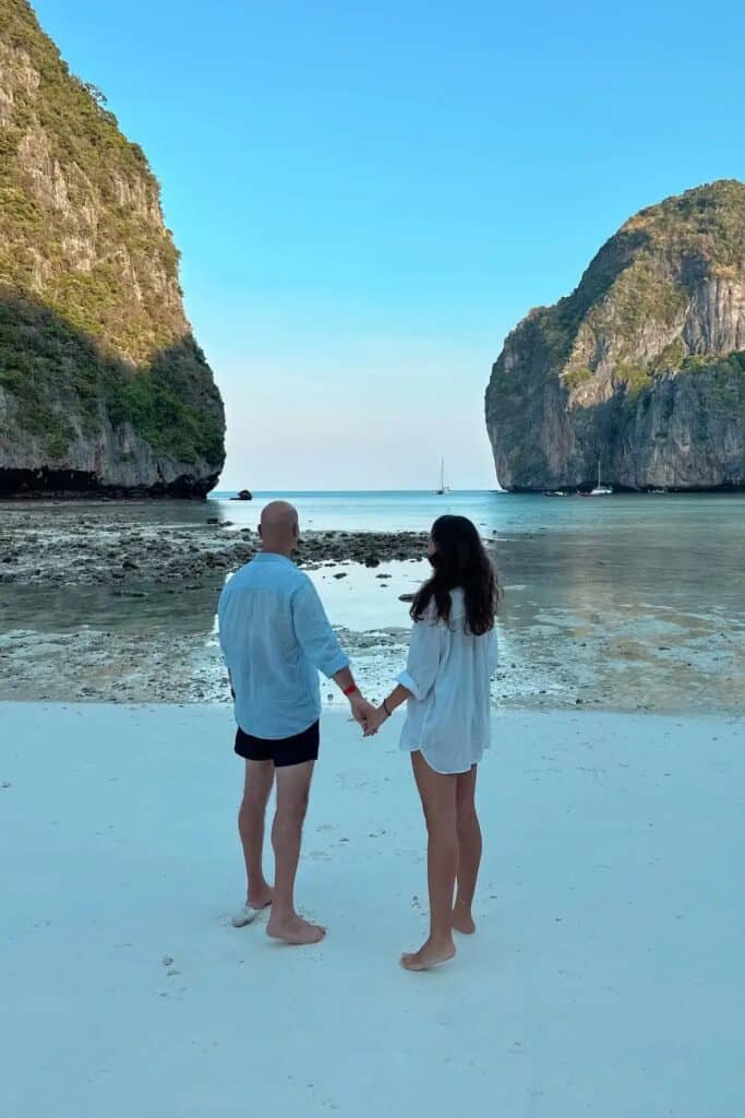 Phi Phi Maya Bay’de beyaz kumsalda el ele duran çift ve arka planda kayalık manzara