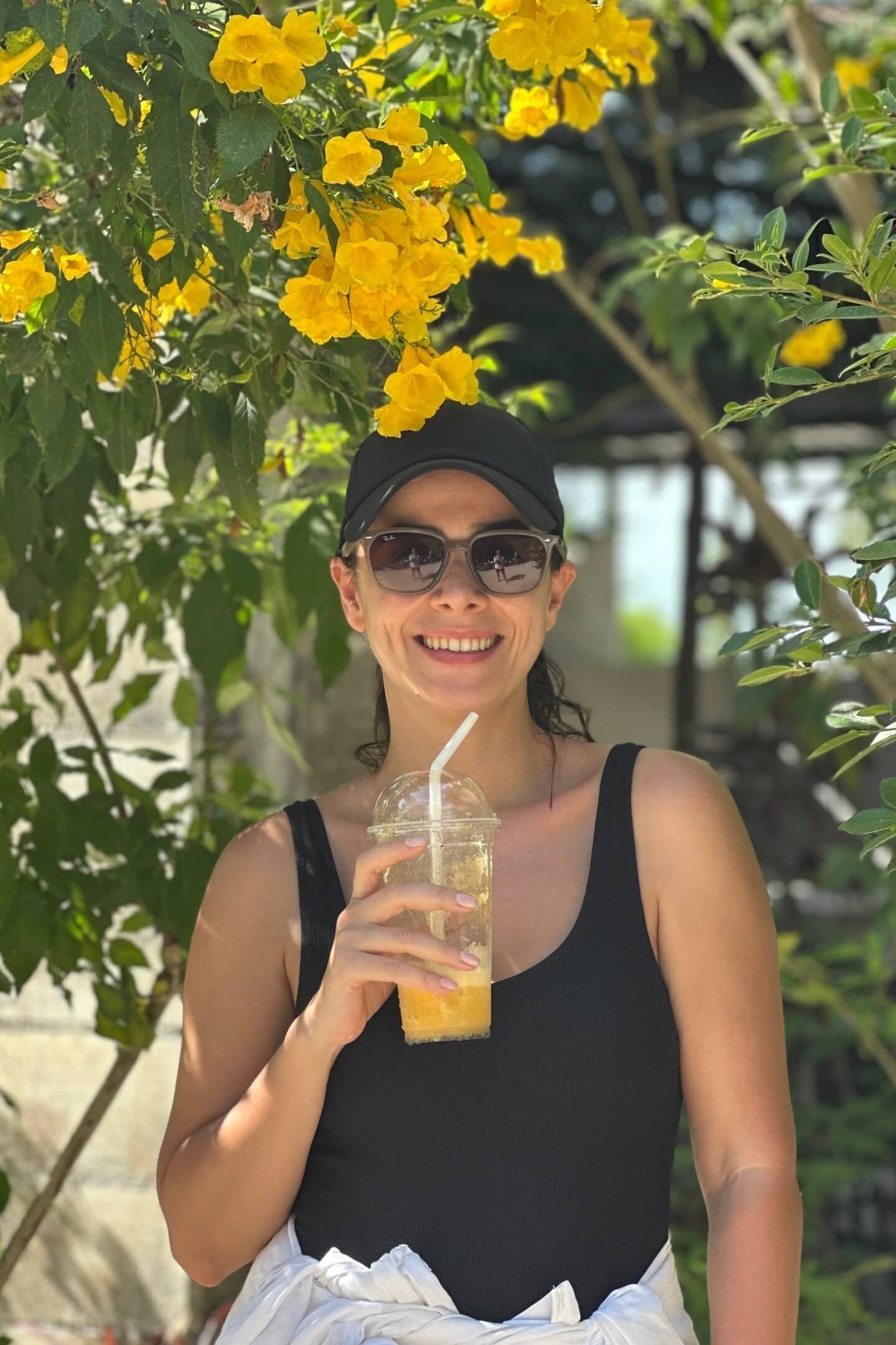 Phuket’te sarı çiçekli bir ağacın altında, elinde soğuk meyve suyu tutan güneş gözlüklü kadın.